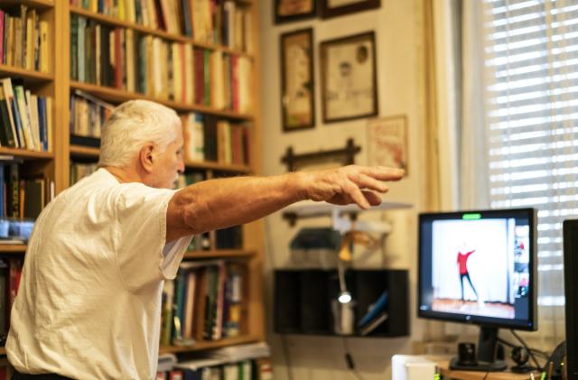 Side view of Senior man, a Parkinson Disease patient, who is standing  in front of monitor, at home, online exercising dancing with help of a physical therapist for Parkinson Disease. Online is only possible therapy as it is Covid-19 lockdown time.