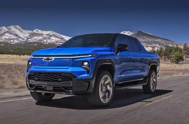 GM marketing photo of the Chevy Silverado EV. The blue truck drives on an open highway with beautiful trees and mountains behind.