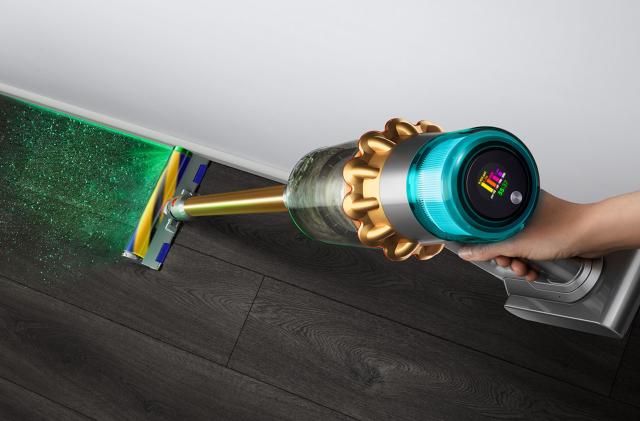 Marketing lifestyle photo of the Dyson V15 Absolute stick vacuum. View from above as a person holds the vacuum. An LCD displays stats on its end, and a green laser path reveals invisible dirt on the floor below.