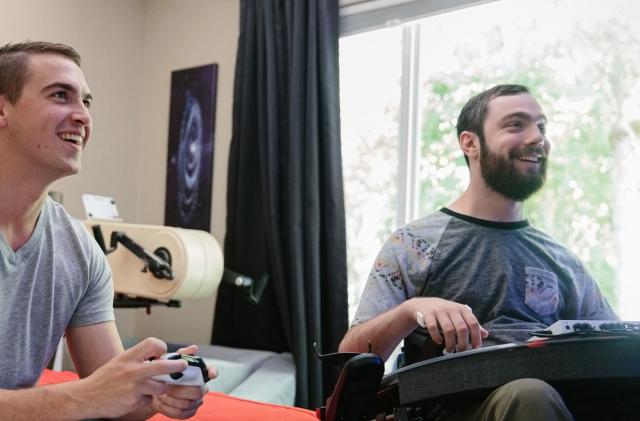 Xbox marketing photo showing two young men playing Xbox games. One of them is using a standard Xbox gamepad, and the other has the Xbox Adaptive Controller.