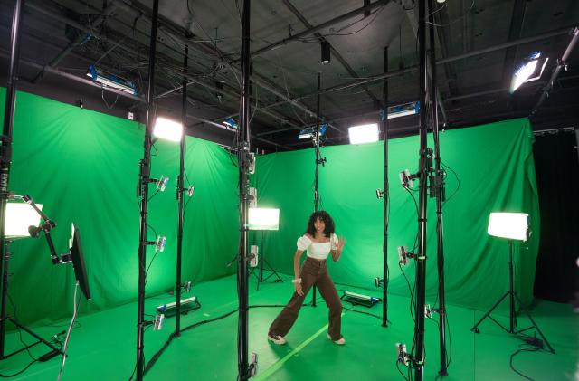 A dancer performs in a volumetric capture stage. Walls are draped with green screens as lights and sensors surround them.