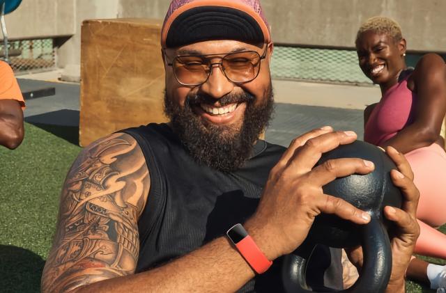 Lifestyle marketing photo of a man holding a small kettlebell in his hand with a (red band) Fitbit Charge 6 on his wrist. A woman sits behind him, stretching, as the two laugh together in an outdoor fitness area.