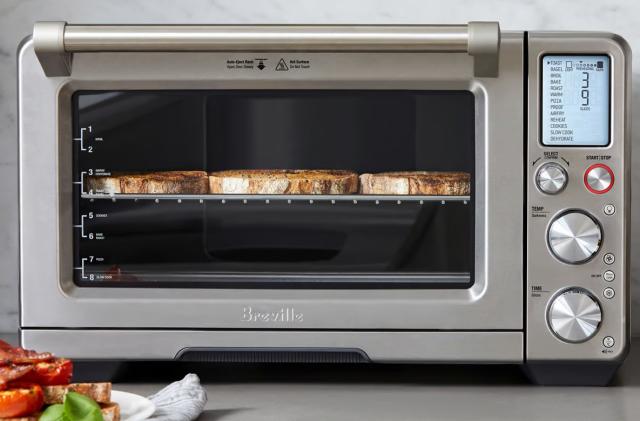Marketing lifestyle photo of the Breville Smart Oven Air Fryer Pro. The oben has at least three slices of thick toast inside (side by side) with sandwiches sitting on the gray marble counter in front of it.