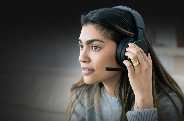 Marketing photo for the Xbox wireless headset. A young woman wears the black over-ear headset with protruding mic. She faces the left with her left hand over the ear cup. She has a calm but determined look on her face.